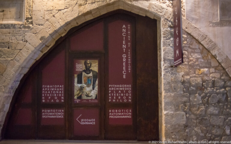 Pointed stone arch framing museum signage about Ancient Greece, Pythagoras, Archimedes, inventions, and robotics.