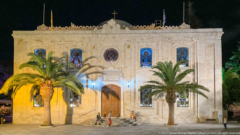 Full night view of Agios Titos Church with palm trees, stained-glass windows, central wooden door, roof cross, and people on the steps.