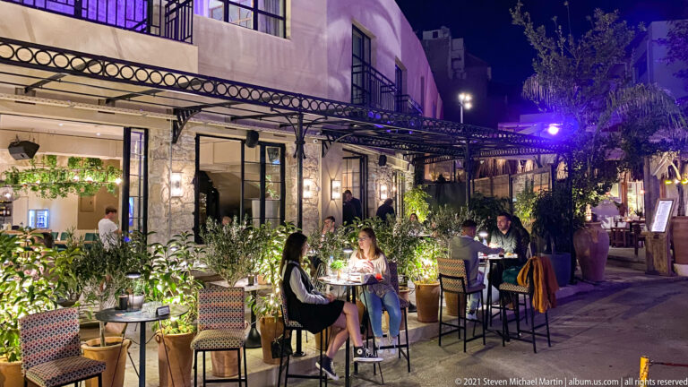 Night restaurant terrace with diners at small tables, potted plants, stone walls, pergola framing, and purple accent lighting.