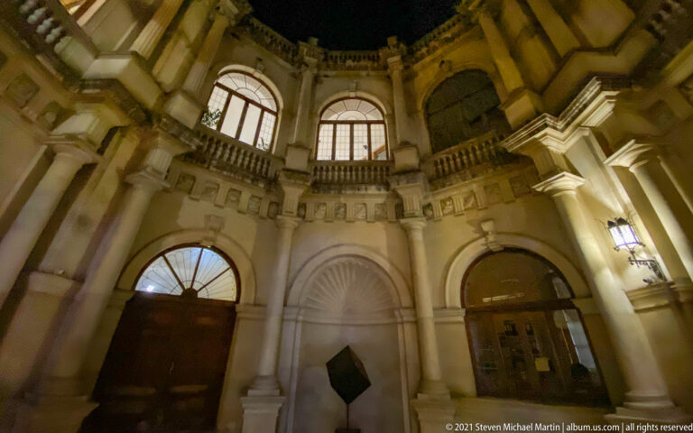 Upward night view inside the Heraklion Loggia with stone arches, shell-headed niche, balustrade, and illuminated upper windows.