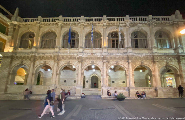 Broad frontal night view of the illuminated Heraklion Loggia with two tiers of arches, ornate balustrade, and pedestrians in front.