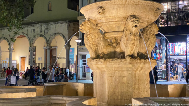 Close night view of carved lion heads beneath the upper basin of the Morosini Fountain, with water streaming into lower basins.