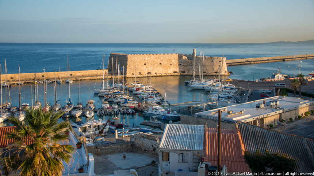 Daylight elevated view of Heraklion marina with sailboats, Koules Fortress, long breakwater, and open Cretan Sea beyond.