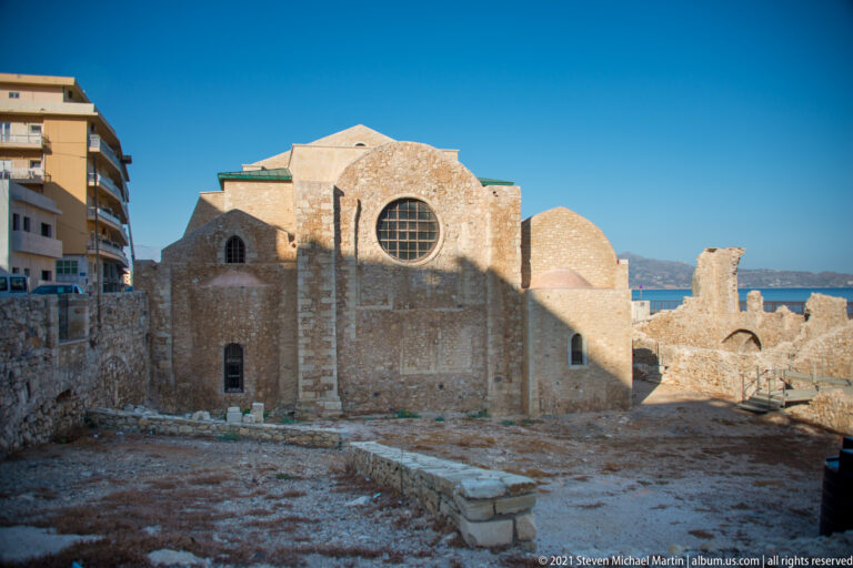 Rear view of Saint Peter of the Dominicans with large circular window, stone walls, excavated foundations, and modern apartments beside it.