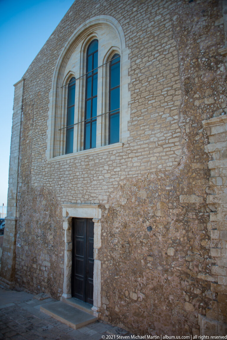 Stone side wall of Saint Peter of the Dominicans with a tall triple-light arched window and small dark doorway.