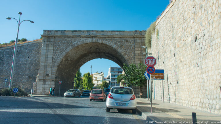 Large stone arch through Heraklion's Venetian walls carrying a roadway, with cars and modern buildings visible beyond.