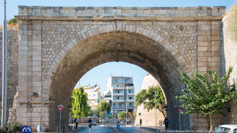 Centered view through a deep stone arch in Heraklion's Venetian walls, framing roadway, trees, and apartment buildings.