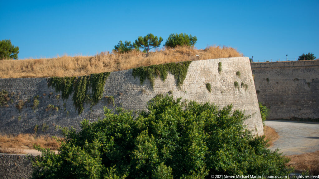 Angular Venetian bastion with sloping stone walls, dry grass, and green vegetation beneath a clear blue sky.