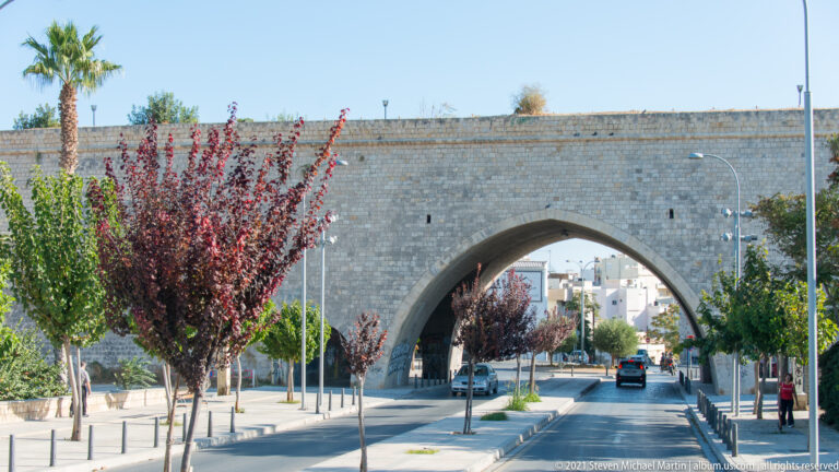 Pointed stone arch through Heraklion's Venetian walls over a modern boulevard lined with trees, streetlights, and cars.
