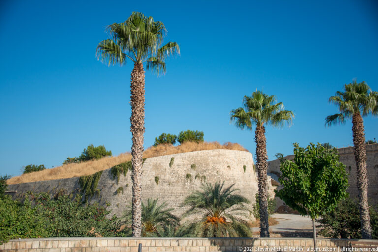 Low sloping Venetian bastion behind palm trees and dense greenery, with dry grass covering the top of the stone wall.
