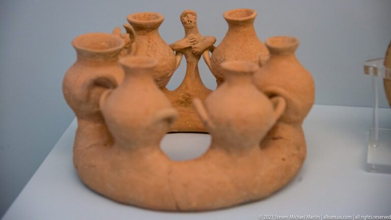 Ring-shaped terracotta object combining several small handled flask vessels around a central standing human figure.