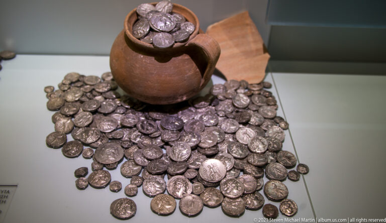 Large spread of ancient silver coins surrounding a small ceramic jug filled with more coins.