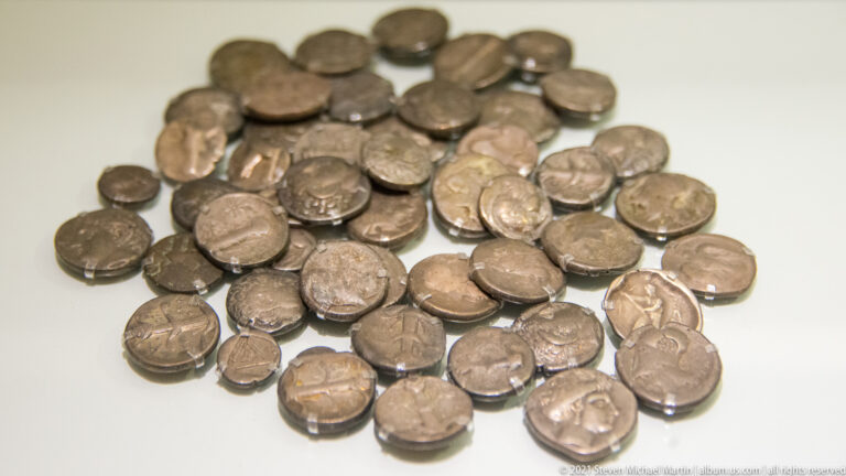 Close view of scattered ancient silver coins showing varied portrait heads, symbols, sizes, and levels of wear.