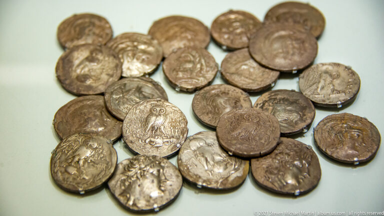 Close-up of ancient silver coins with worn profile portraits, emblematic reverses, irregular edges, and varied relief.