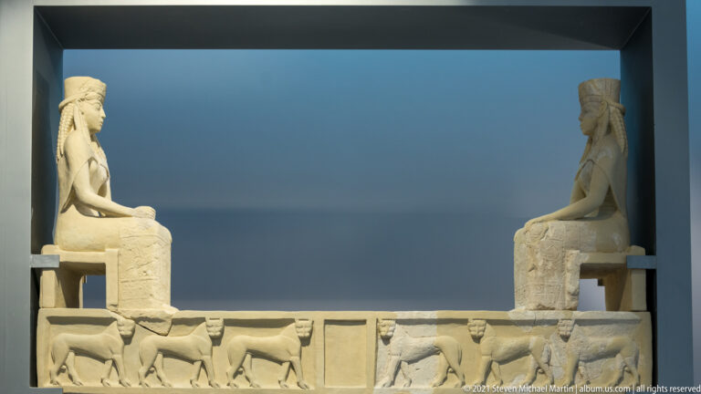 Pale carved stone relief with two seated female figures facing inward above a frieze of walking animals.