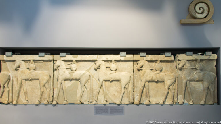 Long pale stone frieze with repeated human figures and horses carved in shallow relief beneath a spiral ornament.