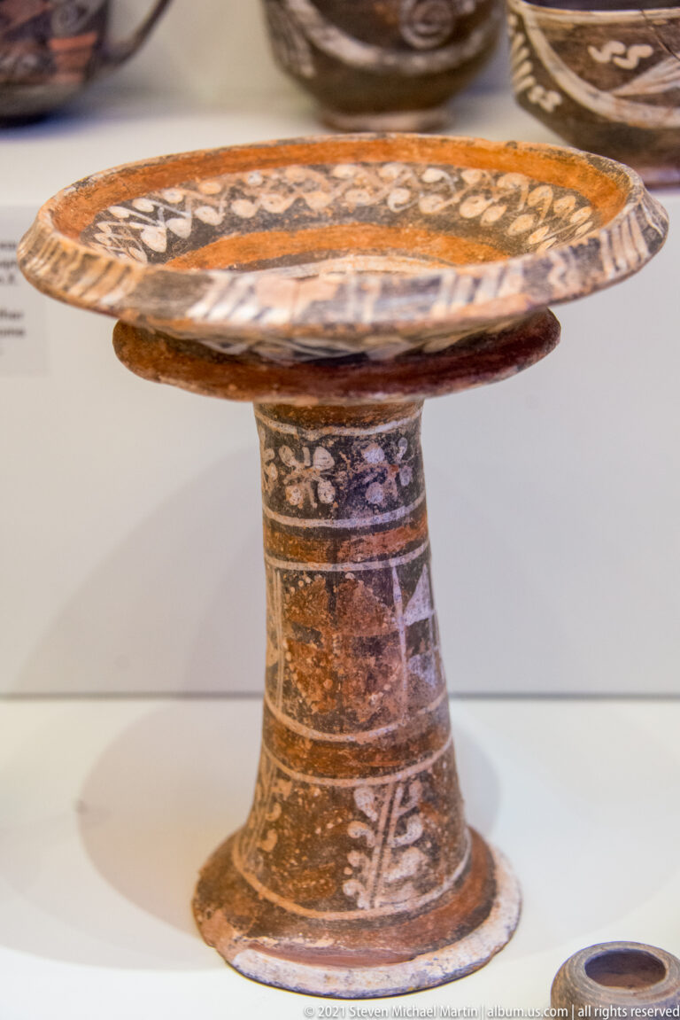 Wide painted ceramic dish on a tall flaring pedestal, decorated with bands and pale rosette-like motifs.