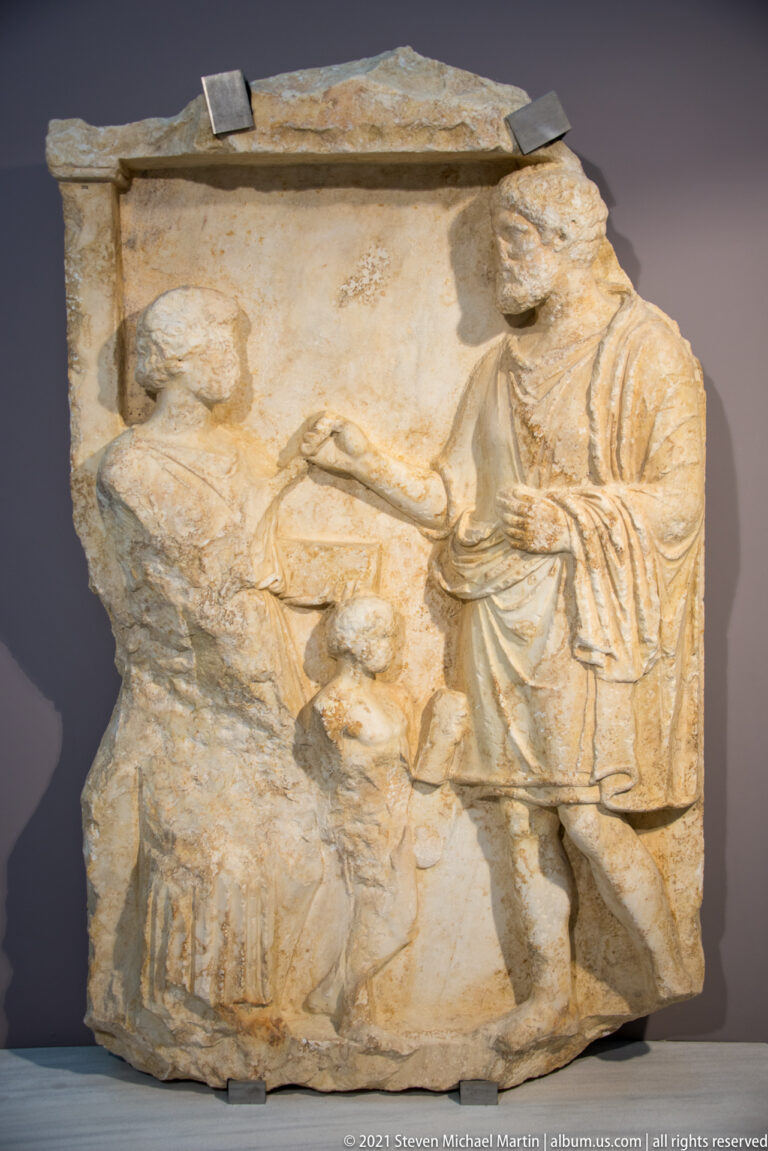 Fragmentary marble relief with a standing draped figure beside a damaged central scene and broken surrounding edges.