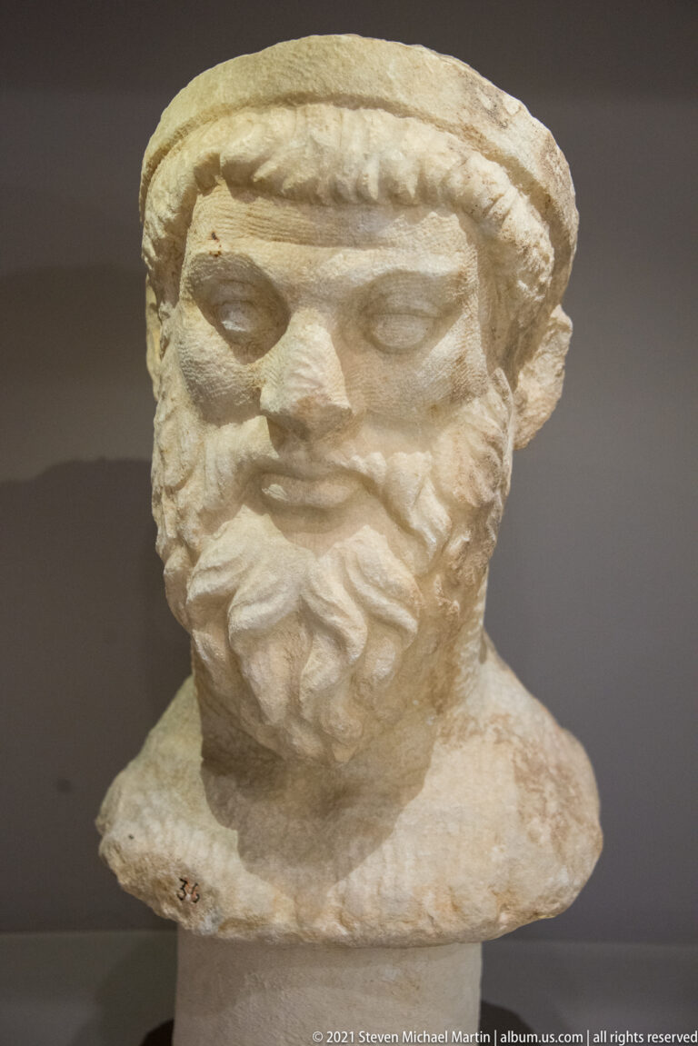 Weathered marble bust of a mature bearded man wearing a low headband or cap.