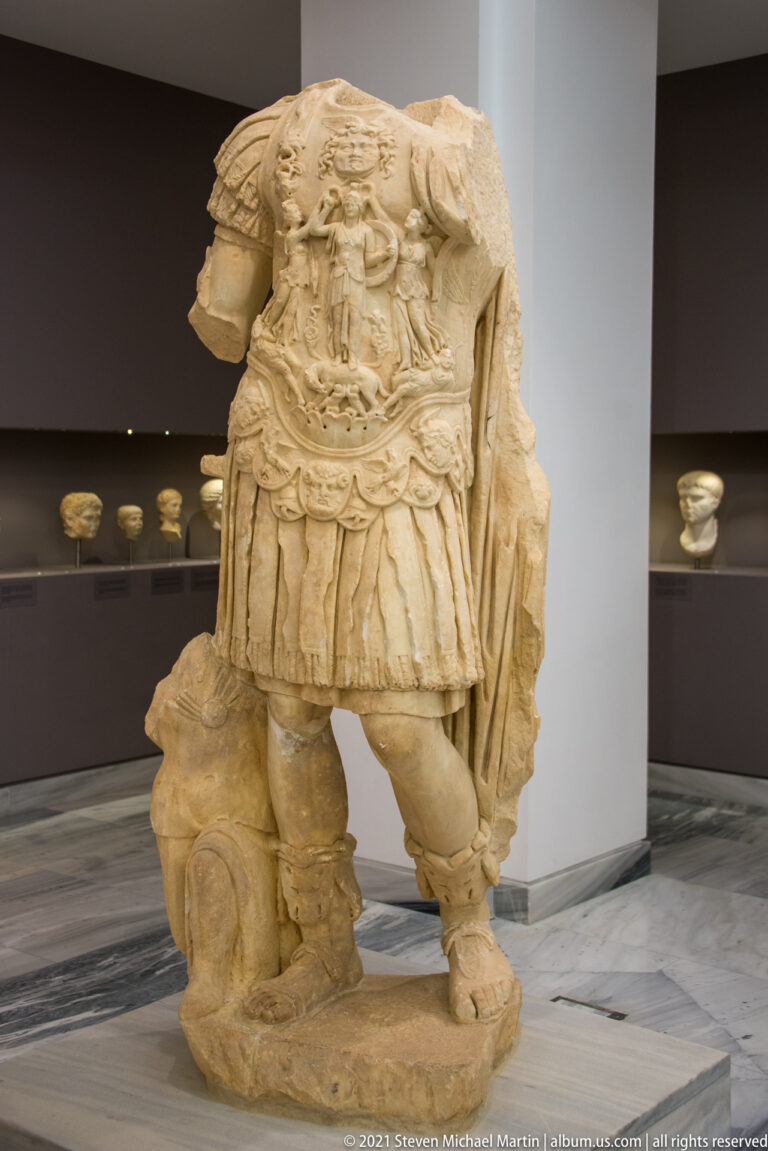 Headless marble military statue wearing an ornate relief-decorated cuirass, short pleated tunic, cloak, and sculpted support.