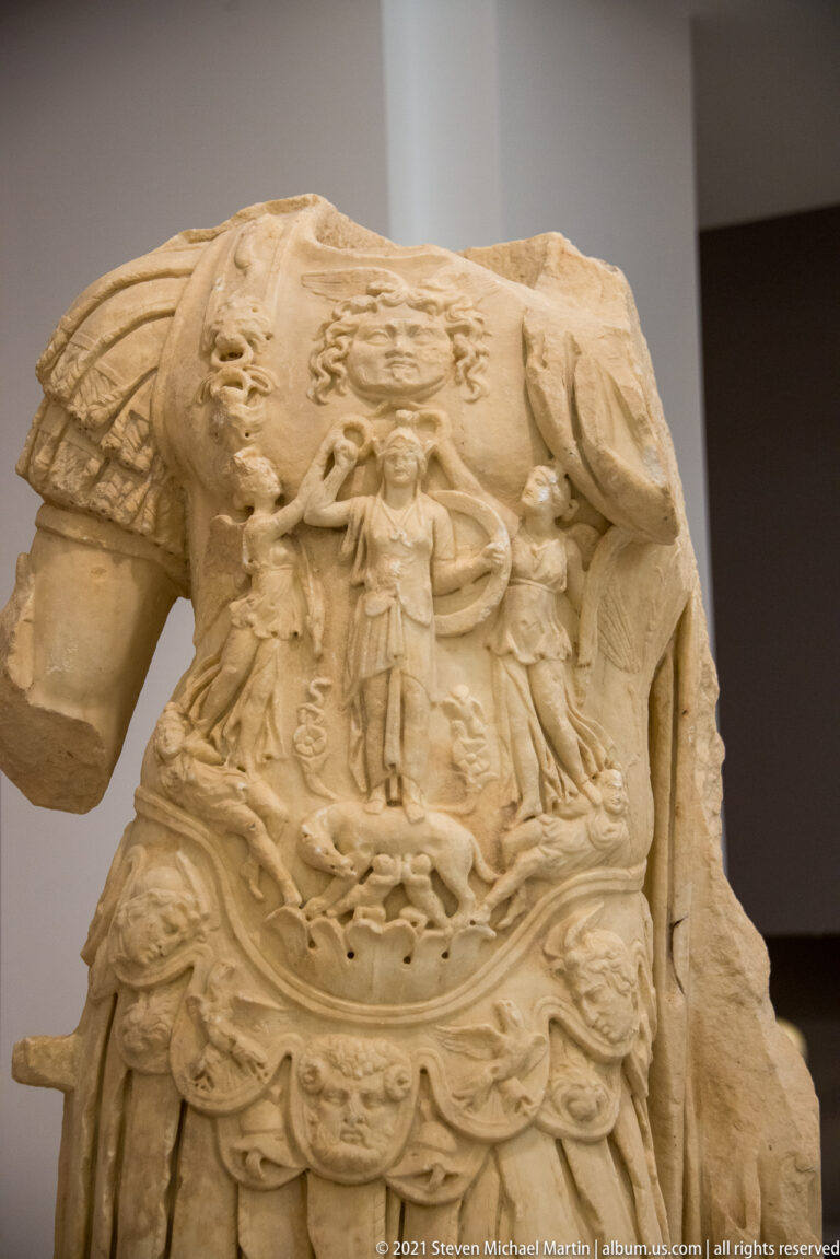 Close view of an ornate marble cuirass carved with central figures, mythological forms, animals, and scrolling ornament.