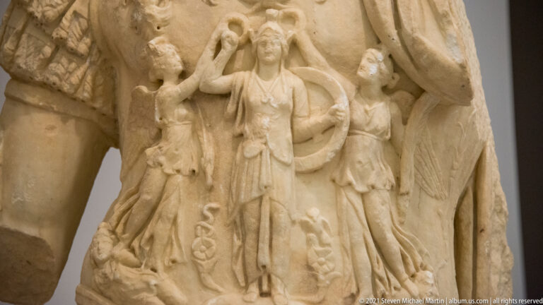 Close-up of two standing female figures carved in relief on the side of a marble military cuirass.