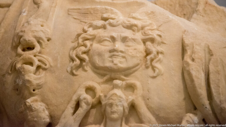 Close-up of a mask-like face with curling hair, likely a gorgoneion, carved on a marble military cuirass.