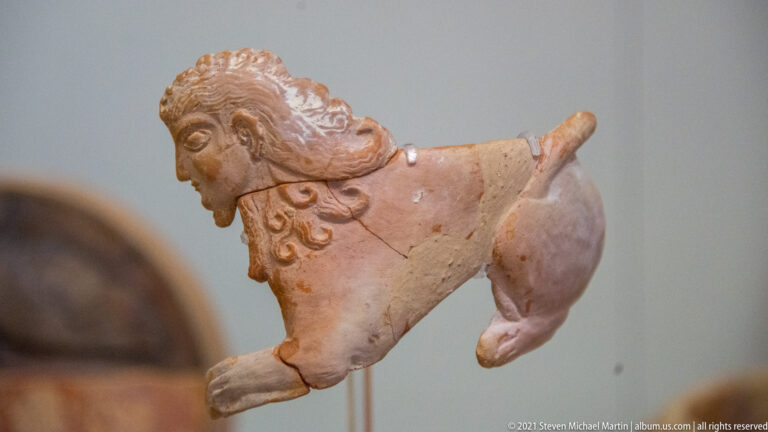 Fragmentary terracotta quadruped with modeled face, curling mane, surviving foreleg, rounded body, and raised tail.