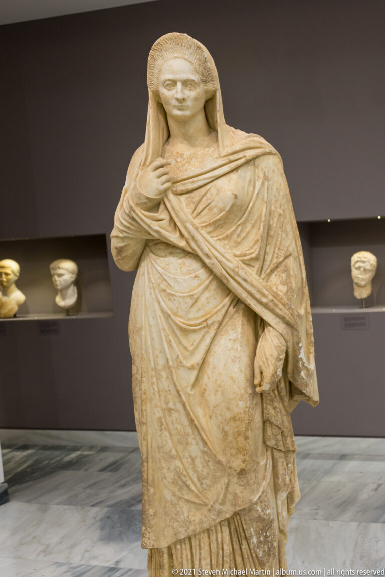 Marble statue of a standing woman wrapped in a heavy mantle, with covered head and one hand raised to her chest.