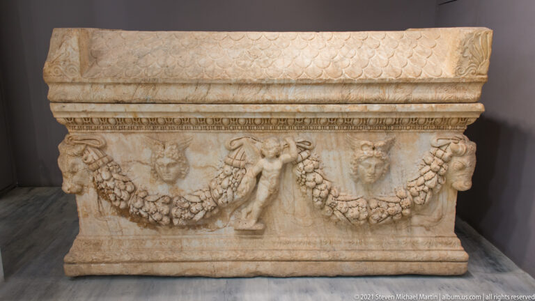 Large marble sarcophagus decorated with hanging garlands, masks, standing figures, and patterned bands along the upper rim.