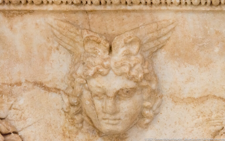 Close-up of a youthful curly-haired head with small wings carved in relief on a marble sarcophagus.