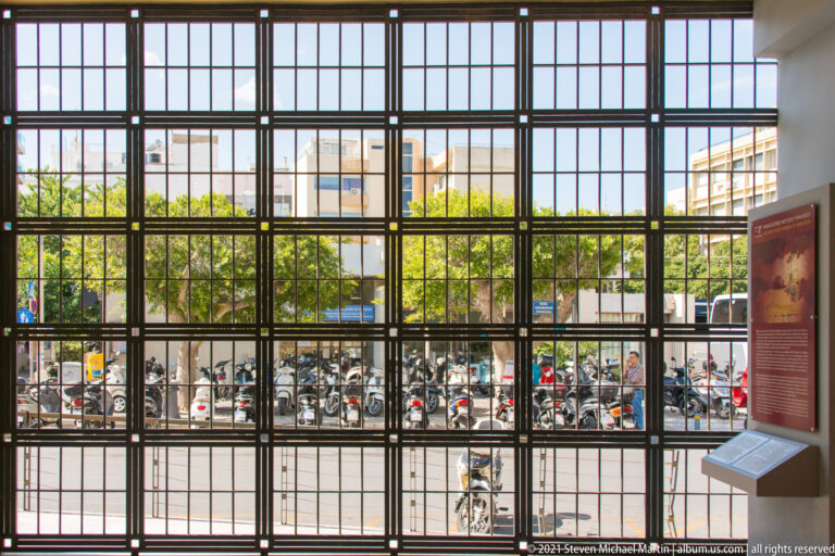 Large black-framed museum window overlooking trees, parked scooters, and city buildings, with an interpretation panel at right.