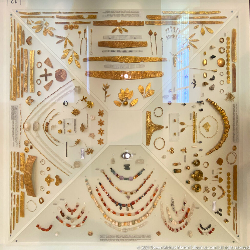 Museum case filled with gold ornaments, necklaces, beads, rings, pendants, pins, and other small archaeological objects.