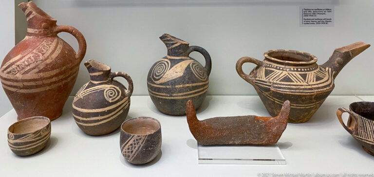 Display of painted jugs and cups with spiral, striped, and geometric decoration, plus a small horned animal-shaped vessel.