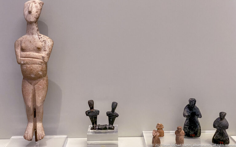 Group of small ancient human figurines in stone, bronze, and terracotta beside a taller fragmentary standing male figure.