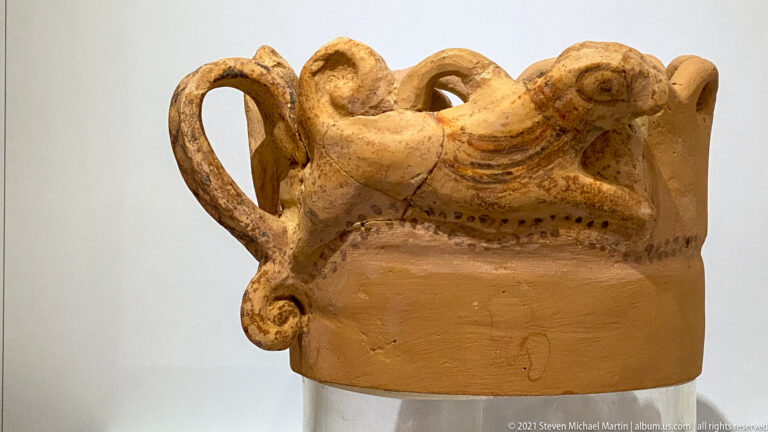 Terracotta vessel fragment with a modeled quadruped across the rim, loop handle, and traces of painted dots and bands.