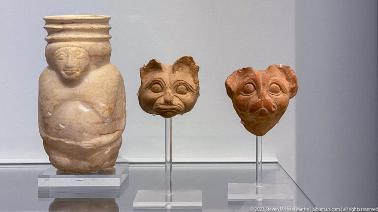 Small pale stone human figure displayed beside two mask-like heads with exaggerated eyes, brows, noses, and mouths.