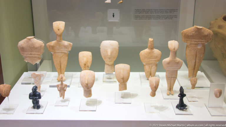 Museum case containing many stylized human figurines, including pale elongated forms and small dark standing figures.