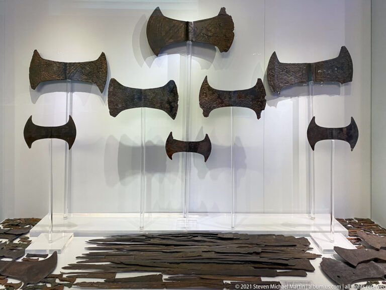 Museum case with several large bronze double axes mounted above rows of long blades and smaller metal implements.