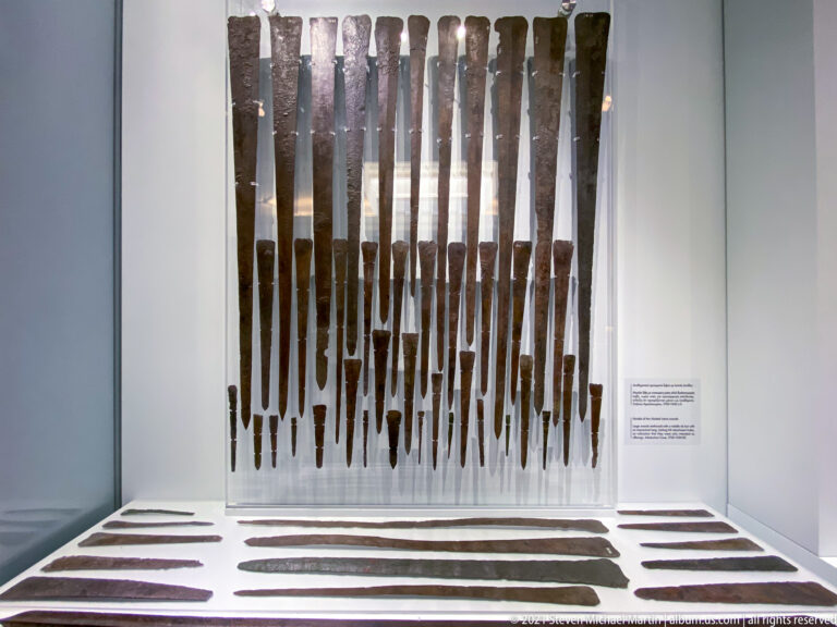 Rows of long patinated bronze swords and blades mounted vertically and horizontally in a museum case.