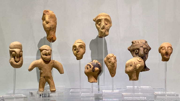 Display of small terracotta human heads and one standing figurine, with varied facial shapes, expressions, and traces of paint.