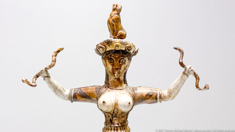 Painted-faience Snake Goddess holding snakes in both hands, with a feline on her head and a flounced skirt.