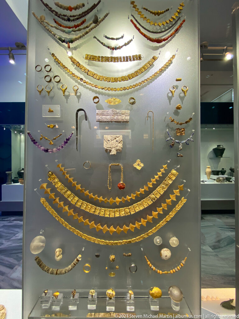 Large museum case of layered necklaces, gold ornaments, colored beads, rings, pendants, pins, and small personal objects.