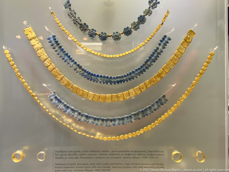 Close display of layered ancient necklaces made from gold beads, rectangular gold plaques, and deep blue beads, with rings below.