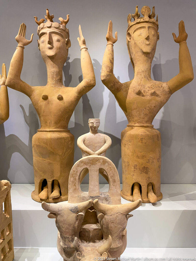 Two large terracotta female figures with raised arms flank a smaller shrine-like model with a central opening and animal-head projections.