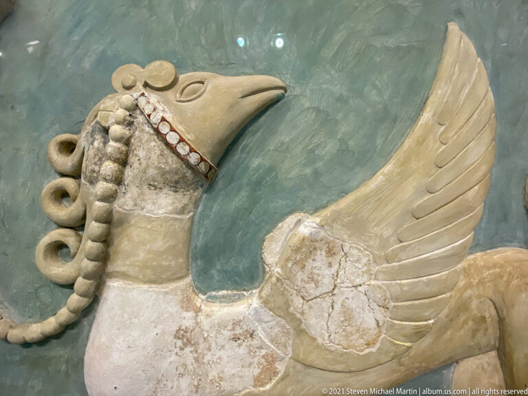 Profile of a Minoan griffin with long beak, curling crest, red-and-white collar, pale body, and large feathered wing.