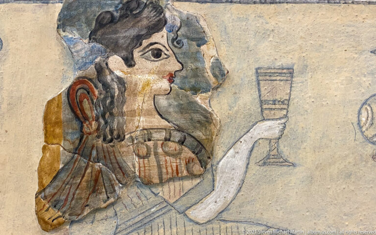 Profile of a seated Minoan woman with dark curling hair and patterned clothing, holding a tall stemmed cup.