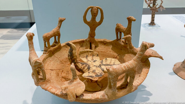 Shallow terracotta circular model populated by long-necked animals and human figures, including a central figure with raised arms.