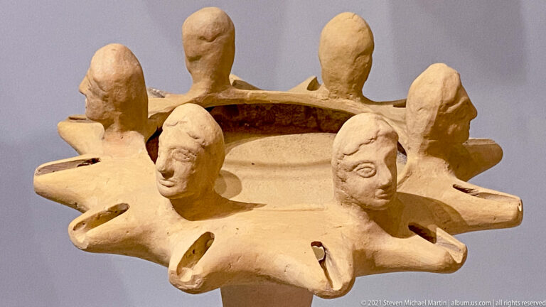 Ring-shaped terracotta vessel encircled by modeled human heads and pointed projections, with taller rounded elements rising behind.