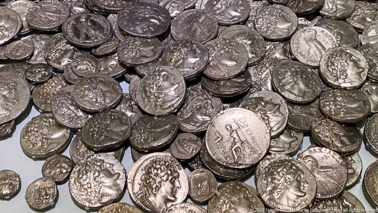 Close view of a dense pile of ancient silver coins with profile portraits, reverse figures, symbols, and irregular edges.